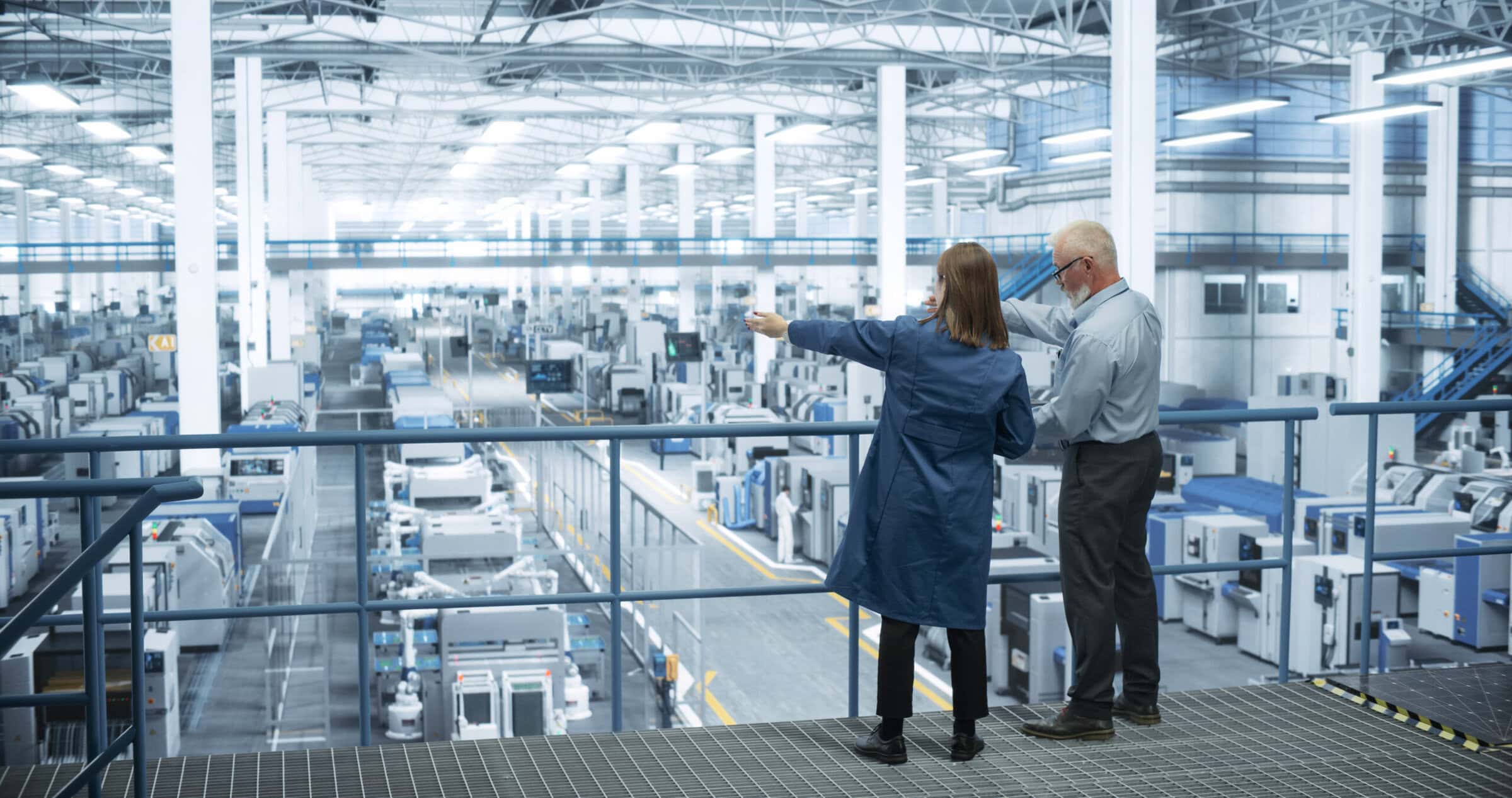 Male and female engineers standing on a platform with their back to the camera and discussing production at an automated electronics manufacturer