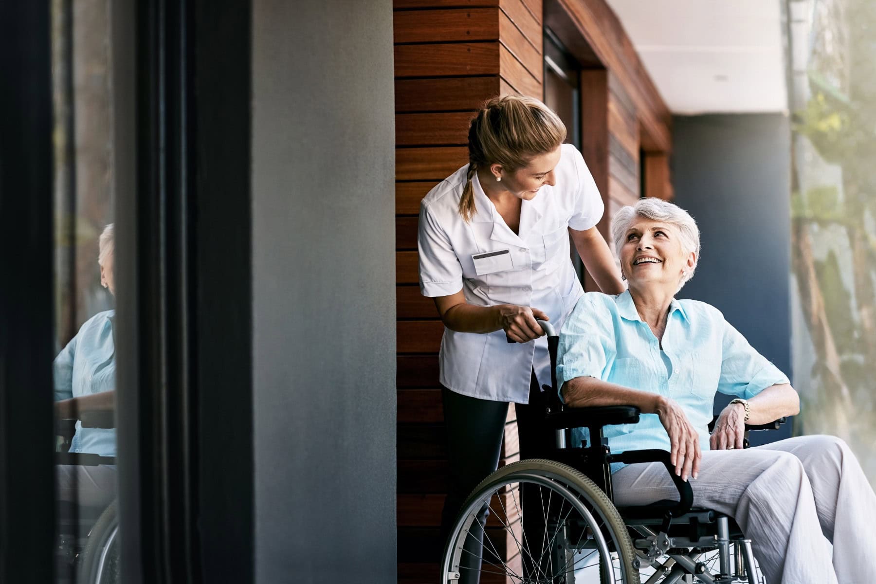 Nurse,,Talking,And,Senior,Woman,On,Wheelchair,For,Support,Or Senior women seated in a wheelchair being pushed by a nurse and smiling
