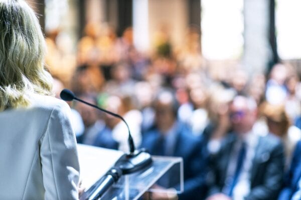 Female,Speaker,At,Business,Conference,,Corporate,Presentation,,Workshop,,Coaching,Training, Professional standing and speaking at a conference